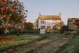 A charming country house with red-framed windows and a white picket fence, surrounded by an autumn landscape. The red and orange leaves create a warm and inviting atmosphere, and the gravel path leads up to the entrance. The house is accompanied by a small stone wall and is set against a clear blue sky.