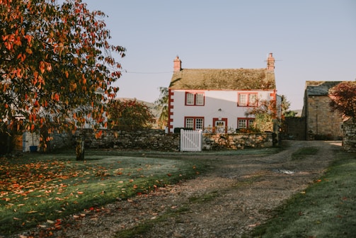 A charming country house with red-framed windows and a white picket fence, surrounded by an autumn landscape. The red and orange leaves create a warm and inviting atmosphere, and the gravel path leads up to the entrance. The house is accompanied by a small stone wall and is set against a clear blue sky.