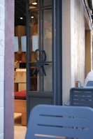 A modern cafe entrance with a glass door featuring a unique decorative symbol. Inside, digital menu boards are visible along with wooden and red upholstered seating. The exterior wall is light-colored and textured, with outdoor seating made of blue metal chairs.