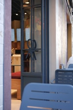 A modern cafe entrance with a glass door featuring a unique decorative symbol. Inside, digital menu boards are visible along with wooden and red upholstered seating. The exterior wall is light-colored and textured, with outdoor seating made of blue metal chairs.