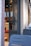 A modern cafe entrance with a glass door featuring a unique decorative symbol. Inside, digital menu boards are visible along with wooden and red upholstered seating. The exterior wall is light-colored and textured, with outdoor seating made of blue metal chairs.