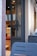 A modern cafe entrance with a glass door featuring a unique decorative symbol. Inside, digital menu boards are visible along with wooden and red upholstered seating. The exterior wall is light-colored and textured, with outdoor seating made of blue metal chairs.