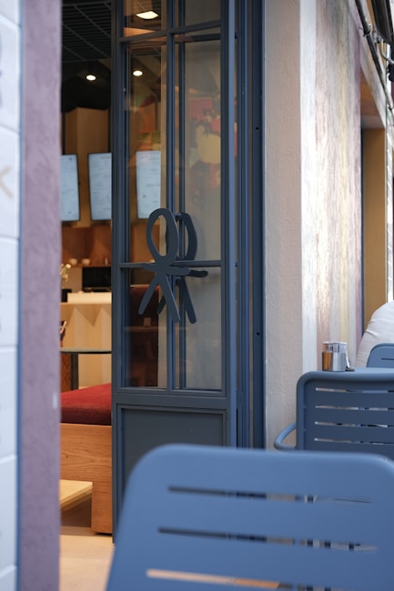 A modern cafe entrance with a glass door featuring a unique decorative symbol. Inside, digital menu boards are visible along with wooden and red upholstered seating. The exterior wall is light-colored and textured, with outdoor seating made of blue metal chairs.