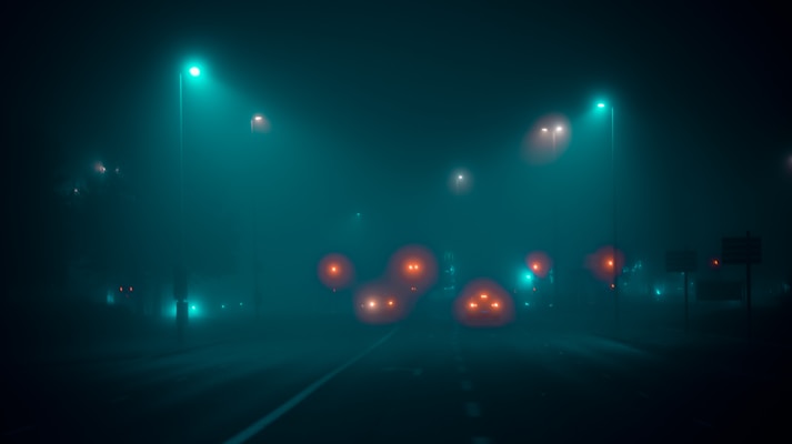 A foggy night scene illuminated by streetlights. The road is bordered by glowing orbs of varying sizes, resembling car headlights obscured by the mist. The atmosphere is eerie and mysterious, with a soft bluish-green tint dominating the scene.