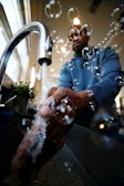 Water flows from a modern chrome faucet as a person washes their hands, surrounded by bubbles. The setting appears to be a kitchen or bathroom with natural light coming through the windows. A plant is visible in the background.