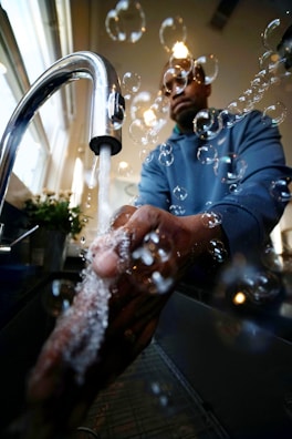 Water flows from a modern chrome faucet as a person washes their hands, surrounded by bubbles. The setting appears to be a kitchen or bathroom with natural light coming through the windows. A plant is visible in the background.