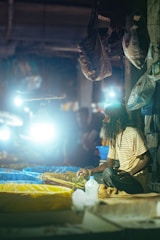 A person with long hair and a beard is sitting on a platform at a market, surrounded by large bags. The area is illuminated with bright artificial lights, creating a glowing effect. The individual is wearing a striped shirt and is accompanied by plastic containers and possibly some goods or produce.
