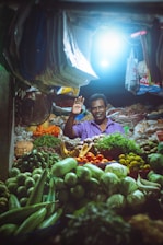 A market vendor stands in a vibrant stall filled with a variety of fresh fruits and vegetables. The stall is illuminated by a bright overhead light, highlighting the colorful produce including green gourds, limes, tomatoes, cucumbers, and leafy greens. The vendor is smiling and waving, creating a welcoming atmosphere.