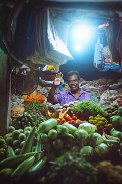 A market vendor stands in a vibrant stall filled with a variety of fresh fruits and vegetables. The stall is illuminated by a bright overhead light, highlighting the colorful produce including green gourds, limes, tomatoes, cucumbers, and leafy greens. The vendor is smiling and waving, creating a welcoming atmosphere.