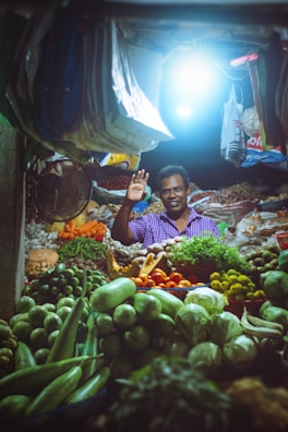 A market vendor stands in a vibrant stall filled with a variety of fresh fruits and vegetables. The stall is illuminated by a bright overhead light, highlighting the colorful produce including green gourds, limes, tomatoes, cucumbers, and leafy greens. The vendor is smiling and waving, creating a welcoming atmosphere.