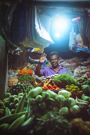 A market vendor stands in a vibrant stall filled with a variety of fresh fruits and vegetables. The stall is illuminated by a bright overhead light, highlighting the colorful produce including green gourds, limes, tomatoes, cucumbers, and leafy greens. The vendor is smiling and waving, creating a welcoming atmosphere.