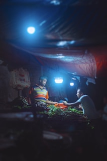 A dimly lit market scene with two people engaged in a transaction. One person, wearing a colorful striped shirt, is positioned behind a table full of fresh vegetables, while the other person, in a light colored shirt, is handing over a payment. The setting is illuminated by bright, blue-tinged artificial lights creating a moody atmosphere.