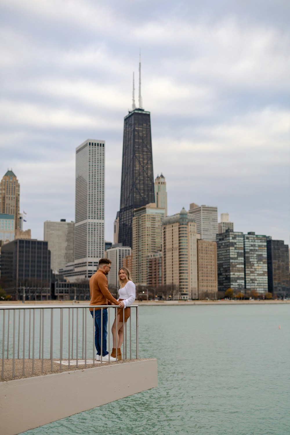 a man and woman posing on a bridge over water with a city in the background