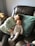A young child and a baby are sitting together on a brown leather sofa. The child is wearing a light-colored sweatshirt and patterned pants, with long hair resting against a light blue pillow. The baby is dressed in a white onesie with tiny patterns, lying comfortably against the child.