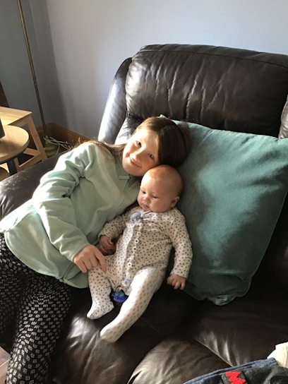 A young child and a baby are sitting together on a brown leather sofa. The child is wearing a light-colored sweatshirt and patterned pants, with long hair resting against a light blue pillow. The baby is dressed in a white onesie with tiny patterns, lying comfortably against the child.