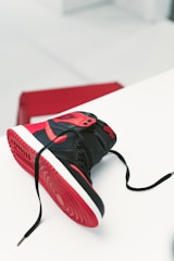 A single black and red high-top sneaker laid on its side with untied laces, displaying its red sole. The sneaker has a prominent logo on the side and is placed on a white surface with a red box visible in the background.