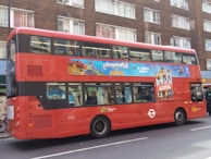 A double-decker red bus in an urban environment, displaying an advertisement for a movie. The bus is parked on a city street, in front of a building with multiple windows.