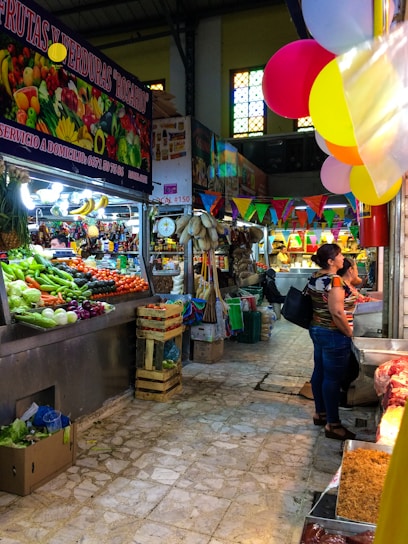 A bustling indoor market scene with various stalls displaying a colorful assortment of fresh fruits and vegetables. Shelves and crates are filled with produce like bananas, onions, and tomatoes. Brightly colored balloons and pennant banners hang throughout, adding a festive atmosphere. Shoppers are seen browsing the goods, and the floor is made of worn, textured tiles.