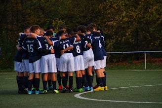 A group of young football players wearing navy blue jerseys and white shorts are huddled together on a green sports field. The players have their arms around each other, suggesting camaraderie and teamwork. The background consists of trees with autumn foliage, indicating an outdoor setting.