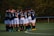 A group of young football players wearing navy blue jerseys and white shorts are huddled together on a green sports field. The players have their arms around each other, suggesting camaraderie and teamwork. The background consists of trees with autumn foliage, indicating an outdoor setting.