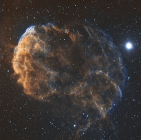 A detailed view of a cosmic nebula with wispy, intricate filaments of gas spanning across the scene, emitting a blend of orange and blue hues. The backdrop is a starry sky, with one particularly bright star standing out prominently to the right of the image.