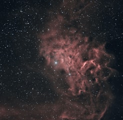 A cosmic scene with a nebula displaying intricate patterns of reddish and pinkish clouds amidst a backdrop of numerous small, sparkling stars. The nebula forms ethereal, smoke-like trails that blend into the dark, expansive space surrounding it.