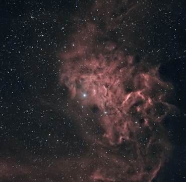 A cosmic scene with a nebula displaying intricate patterns of reddish and pinkish clouds amidst a backdrop of numerous small, sparkling stars. The nebula forms ethereal, smoke-like trails that blend into the dark, expansive space surrounding it.