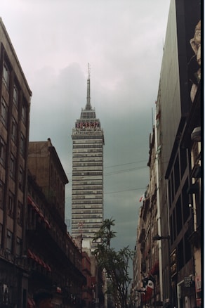 A tall, prominent skyscraper named 'Latinoamericana' rises into a cloudy sky. The building is surrounded by older, shorter buildings on either side of a narrow street. Signs, streetlights, and a few trees are visible along the street, which is busy with pedestrian activity. The overall scene is urban with a mix of modern and historic architecture.