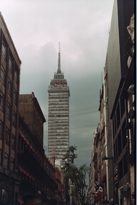 A tall, prominent skyscraper named 'Latinoamericana' rises into a cloudy sky. The building is surrounded by older, shorter buildings on either side of a narrow street. Signs, streetlights, and a few trees are visible along the street, which is busy with pedestrian activity. The overall scene is urban with a mix of modern and historic architecture.