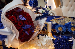 A luxurious arrangement featuring a bouquet of red roses adorned with blue butterflies, set alongside a richly decorated blue and gold-themed table. The setting includes elegant glassware, intricate floral decorations, and a plush white armchair in the background.