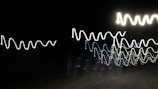 Bright, wavy lines of light against a pitch-black background create an abstract pattern, reminiscent of oscillating waves or electrical signals.