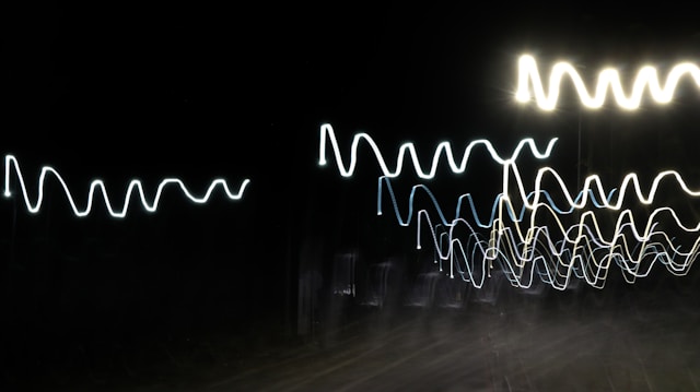 Bright, wavy lines of light against a pitch-black background create an abstract pattern, reminiscent of oscillating waves or electrical signals.