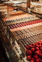 A display case filled with a variety of colorful and neatly arranged pastries or sweets. Each row features different types of confections, including chocolates and round, red truffles. The background includes trays of more pastries, showcasing a rich assortment of treats.