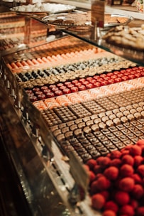 A display case filled with a variety of colorful and neatly arranged pastries or sweets. Each row features different types of confections, including chocolates and round, red truffles. The background includes trays of more pastries, showcasing a rich assortment of treats.
