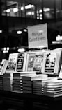 A black and white image features a bookstore shelf labeled 'History & Current Events', showcasing a selection of books. Notable titles include works about Anne Frank, the Battle of the Atlantic, and Auschwitz. The background is out of focus, creating a serene environment typical of a bookstore.
