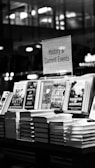 A black and white image features a bookstore shelf labeled 'History & Current Events', showcasing a selection of books. Notable titles include works about Anne Frank, the Battle of the Atlantic, and Auschwitz. The background is out of focus, creating a serene environment typical of a bookstore.