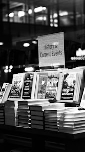A black and white image features a bookstore shelf labeled 'History & Current Events', showcasing a selection of books. Notable titles include works about Anne Frank, the Battle of the Atlantic, and Auschwitz. The background is out of focus, creating a serene environment typical of a bookstore.