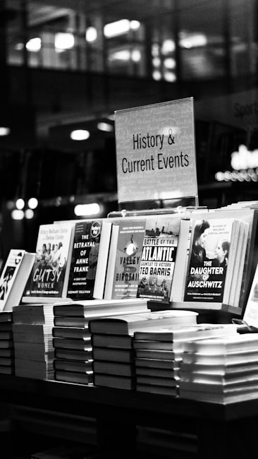 A black and white image features a bookstore shelf labeled 'History & Current Events', showcasing a selection of books. Notable titles include works about Anne Frank, the Battle of the Atlantic, and Auschwitz. The background is out of focus, creating a serene environment typical of a bookstore.