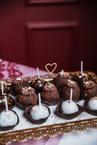 An assortment of decorative cake pops arranged on an ornate golden tray. Some are coated in rich, dark chocolate and topped with sprinkles, while others are covered in white icing with colorful sprinkles. A delicate heart-shaped topper stands out among the treats.