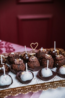 An assortment of decorative cake pops arranged on an ornate golden tray. Some are coated in rich, dark chocolate and topped with sprinkles, while others are covered in white icing with colorful sprinkles. A delicate heart-shaped topper stands out among the treats.