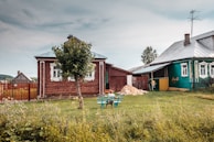 A rural scene with two wooden houses, one painted red and the other green, adorned with decorative window frames. A tree stands in the garden area alongside an empty bench. The sky is overcast, giving a calm and serene atmosphere to the countryside setting. A small pile of sand is visible near the fence, suggesting recent construction or maintenance. Lush green grass and a variety of wildflowers add a touch of color to the foreground.