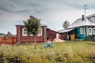 A rural scene with two wooden houses, one painted red and the other green, adorned with decorative window frames. A tree stands in the garden area alongside an empty bench. The sky is overcast, giving a calm and serene atmosphere to the countryside setting. A small pile of sand is visible near the fence, suggesting recent construction or maintenance. Lush green grass and a variety of wildflowers add a touch of color to the foreground.