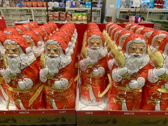 Rows of foil-wrapped chocolate figures resembling Santa Claus are neatly arranged on display in a store. Each figure is decorated with a red outfit, white beard, and festive golden accents. Shelves in the background hold various products, indicating a retail environment.