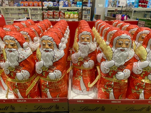 Rows of foil-wrapped chocolate figures resembling Santa Claus are neatly arranged on display in a store. Each figure is decorated with a red outfit, white beard, and festive golden accents. Shelves in the background hold various products, indicating a retail environment.