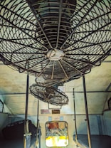 Three ceiling fans are mounted inside a train compartment, showcasing metal grilles and aging infrastructure. The compartment has a worn and industrial look, with a small window and seating area visible below.