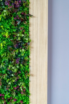 A vertical garden design featuring a lush display of various green plants with a mix of small pink and purple flowers, set against a wooden panel. The arrangement creates a vibrant mix of textures and colors with a natural aesthetic.