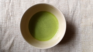 A bowl filled with a frothy green liquid, likely matcha tea, resting on a textured beige fabric surface.