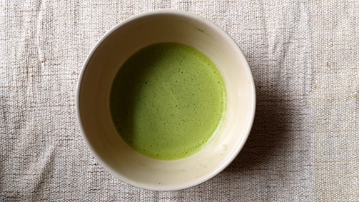 A bowl filled with a frothy green liquid, likely matcha tea, resting on a textured beige fabric surface.