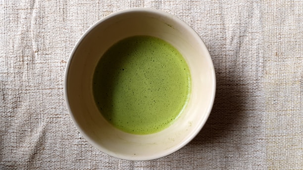 A bowl filled with a frothy green liquid, likely matcha tea, resting on a textured beige fabric surface.