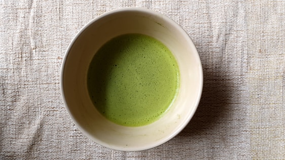 A bowl filled with a frothy green liquid, likely matcha tea, resting on a textured beige fabric surface.